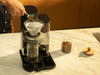 Scooping ground coffee into the glass dripper of a Cosori Juni automatic pour-over coffee machine on a marble counter