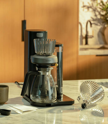 COSORI Juni automatic pour-over coffee maker on a kitchen countertop with cone brew basket, carafe, and coffee accessories