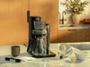 COSORI Juni automatic pour-over coffee maker on a kitchen countertop with cone brew basket, carafe, and coffee accessories