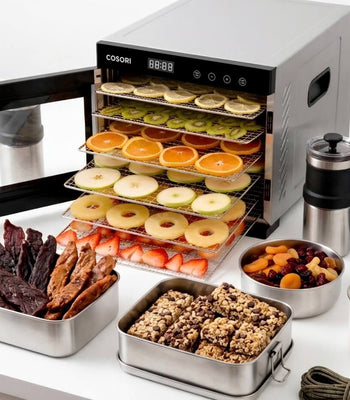 A Cosori food dehydrator filled with sliced fruits next to outdoor camping gear, beef jerky, and homemade granola bars