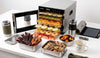 A Cosori food dehydrator filled with sliced fruits next to outdoor camping gear, beef jerky, and homemade granola bars